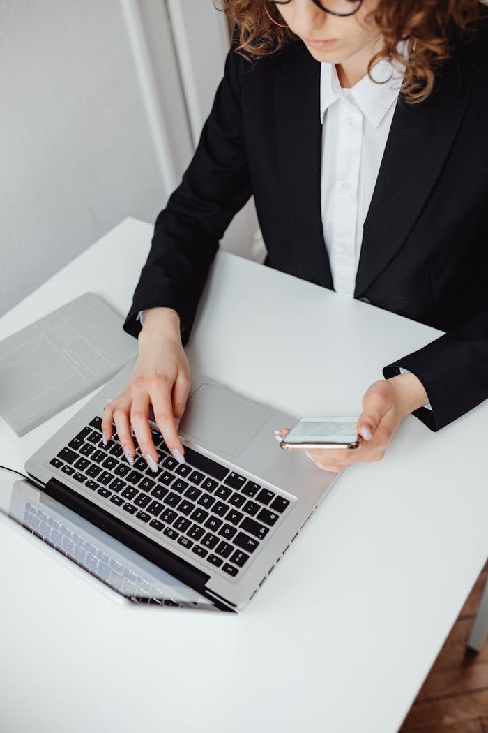 Focused businesswoman in black blazer using laptop and smartphone at desk.